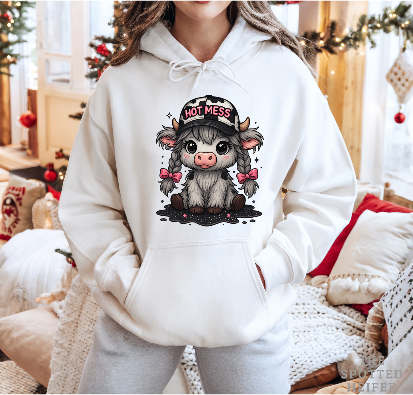 Woman wearing white Hot Mess cow hoodie with pink bows and braids, cozy holiday background, The Spotted Heifer.