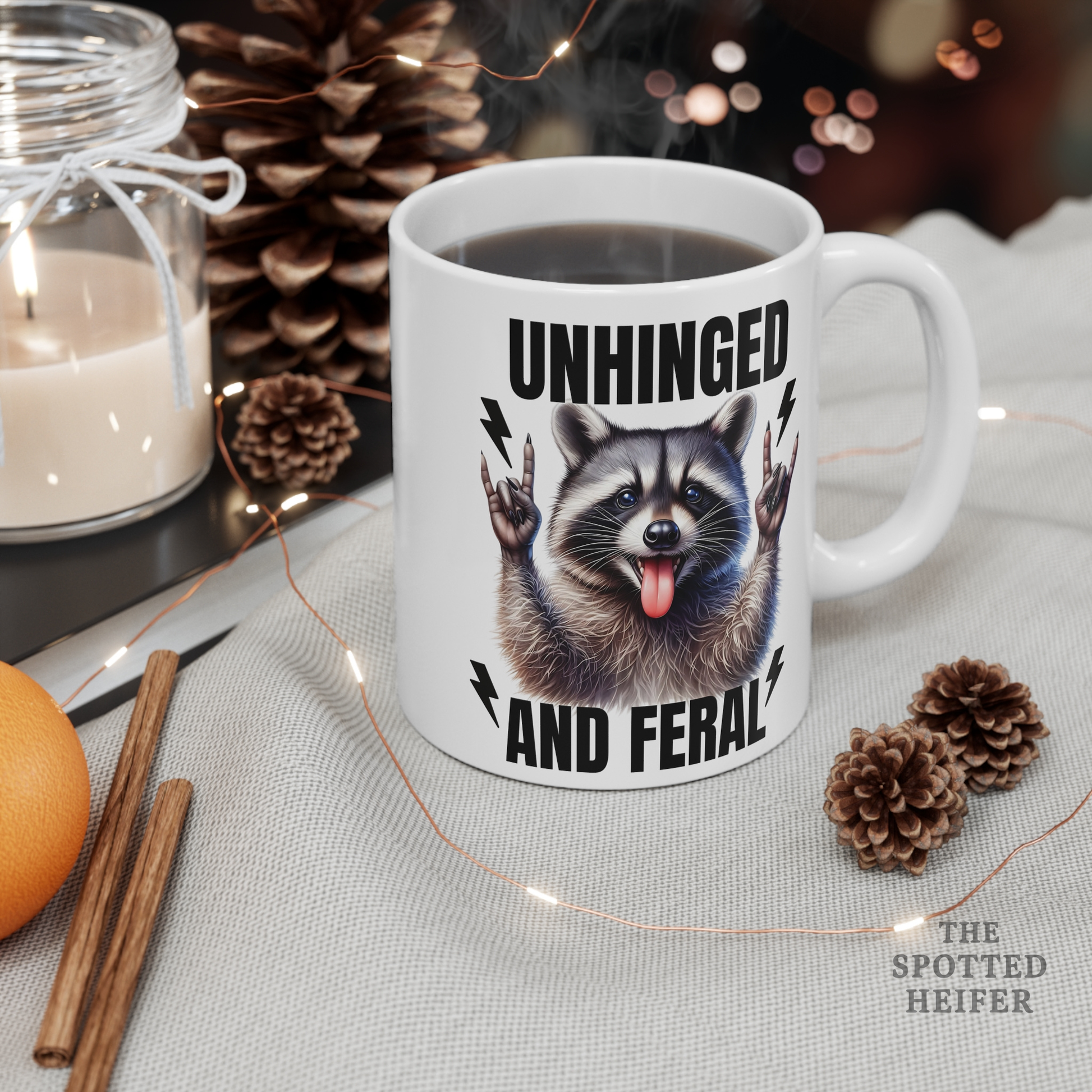 White Unhinged and Feral raccoon coffee mug sitting on a wooden table beside pinecones and coffee beans, cozy fall-style scene.