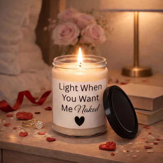Lit soy candle with “Light When You Want Me Naked” label glowing on a cozy table with warm romantic lighting.