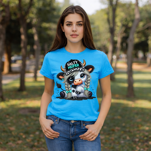 Model wearing Tropical Blue Gildan Softstyle t-shirt featuring a cute cow with big eyes, messy hair, and “Salty Heifer” hat outdoors.