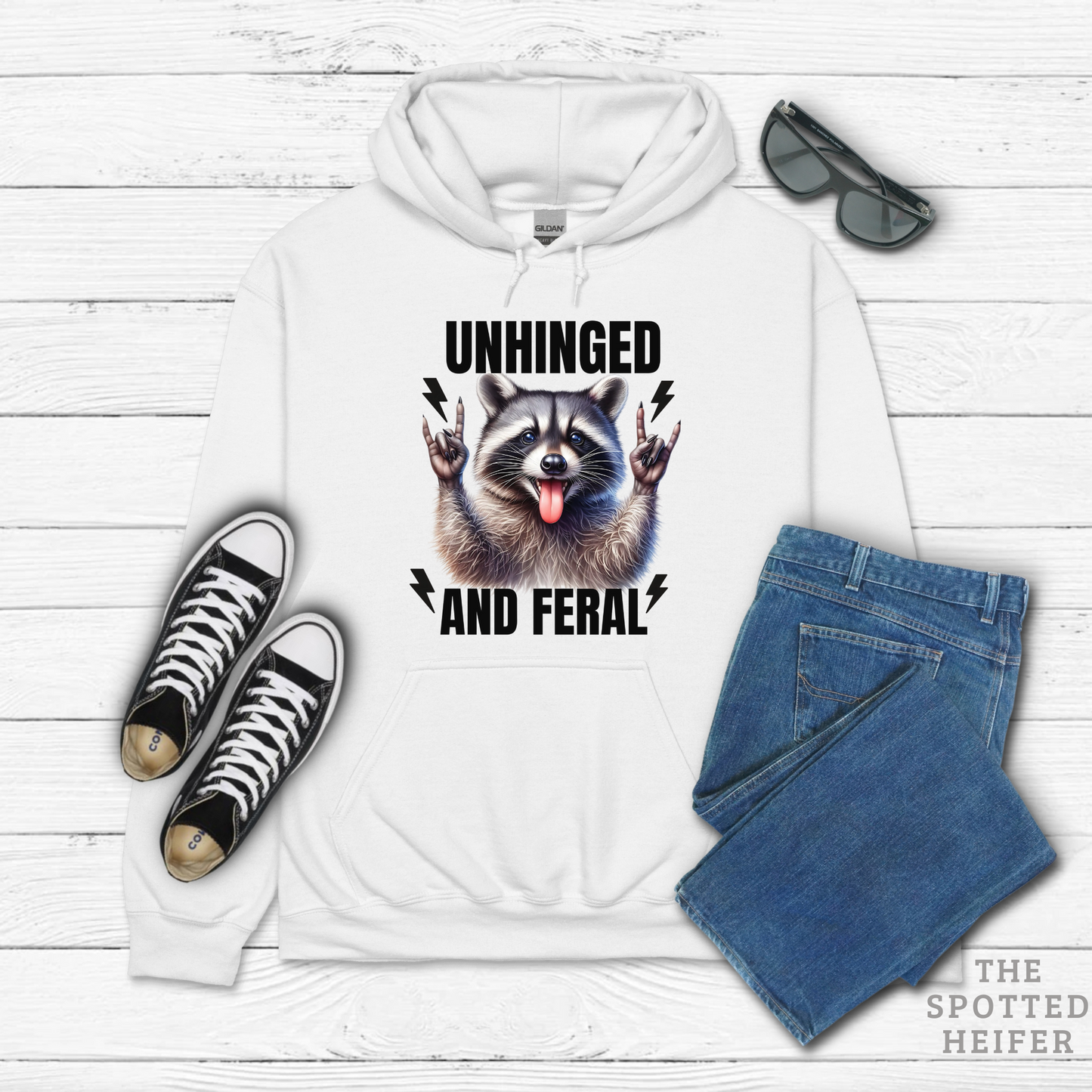 White Gildan hoodie featuring a playful raccoon graphic with the text 'Unhinged and Feral' on the front, showing the raccoon with a mischievous expression and vibrant details.