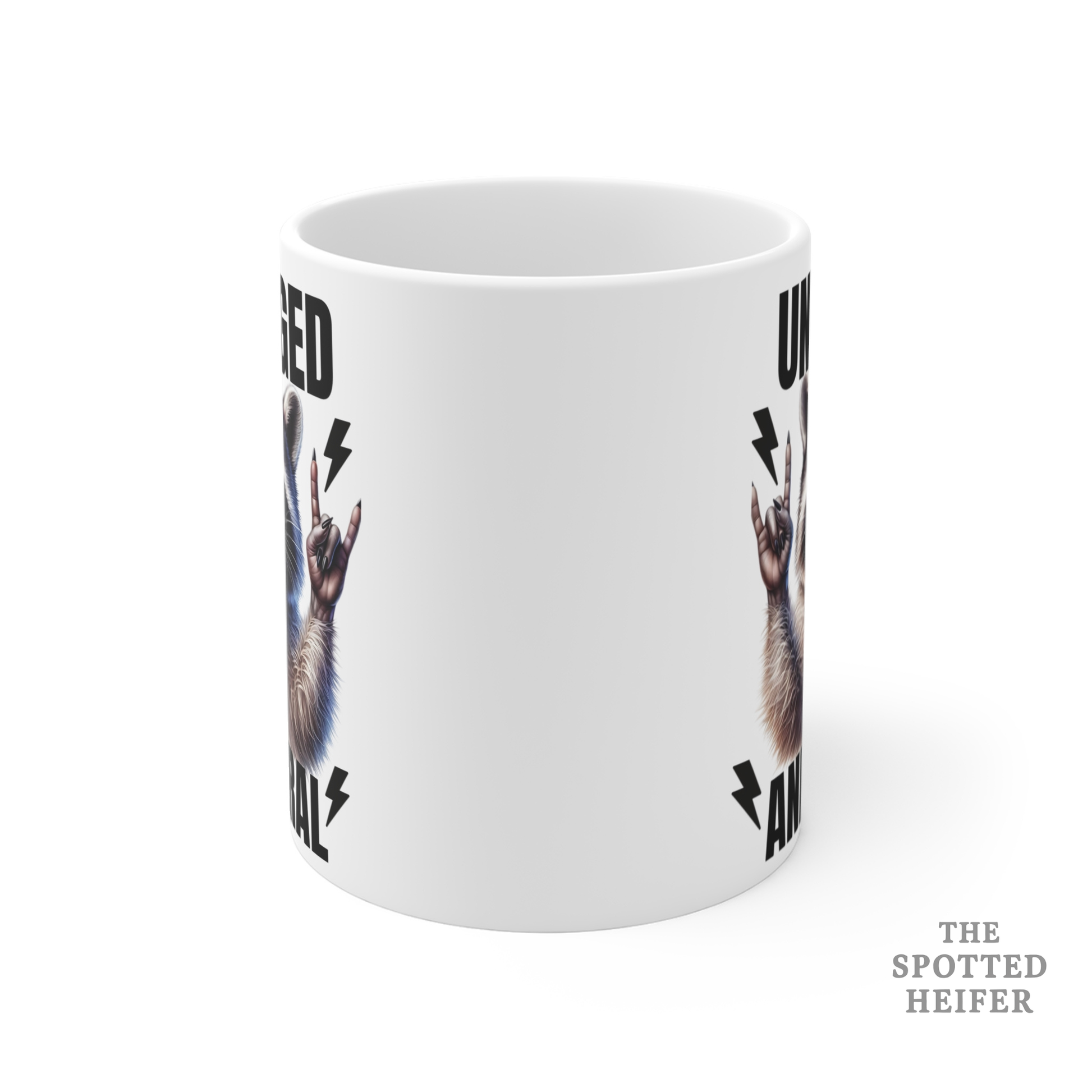 Front view of a white ceramic mug featuring a raccoon graphic with tongue out and hands raised, text reads “Unhinged and Feral,” from The Spotted Heifer.